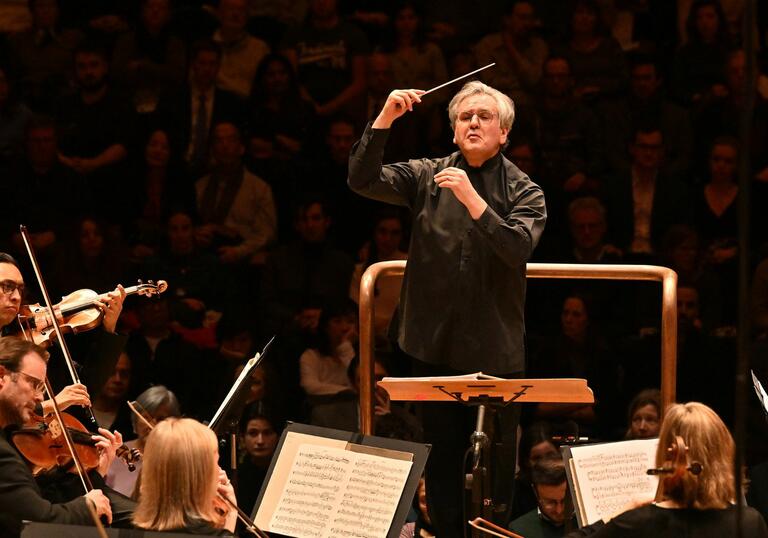 Sir Antonio Pappano conducting the LSO on the Barbican stage, holding a baton, with LSO string players in the foreground of the image and audience members in the background.