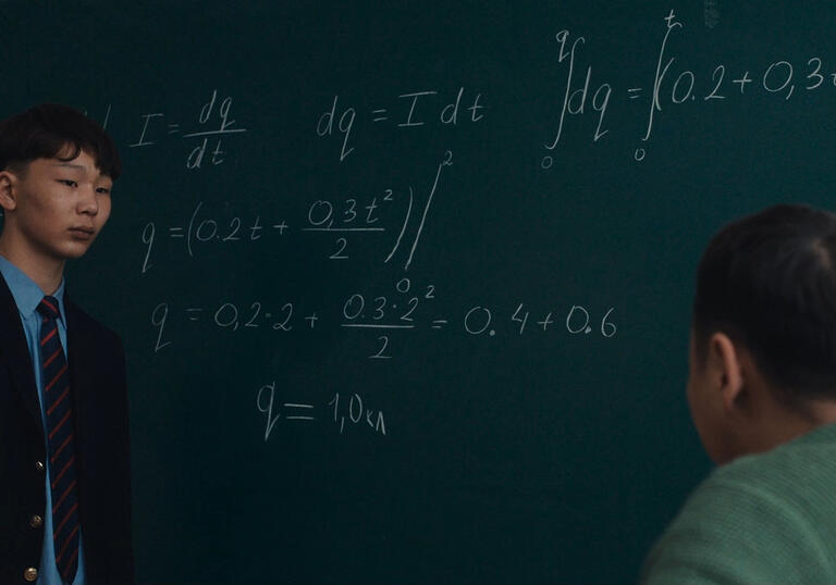 A young boy stands at a school chalkboard in front of his teacher, with complicated mathematics written in chalk.