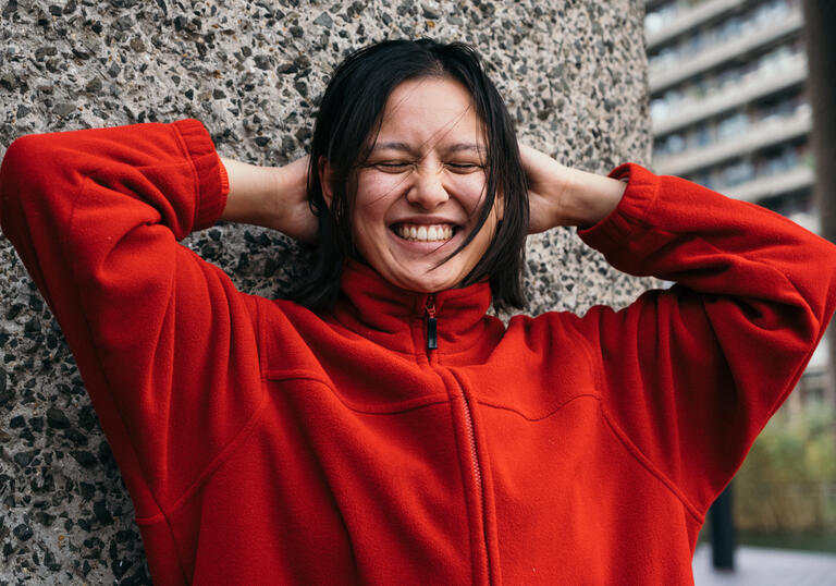 A young poet smiling with hands behind head. Leaning against concrete wall, wearing red top.