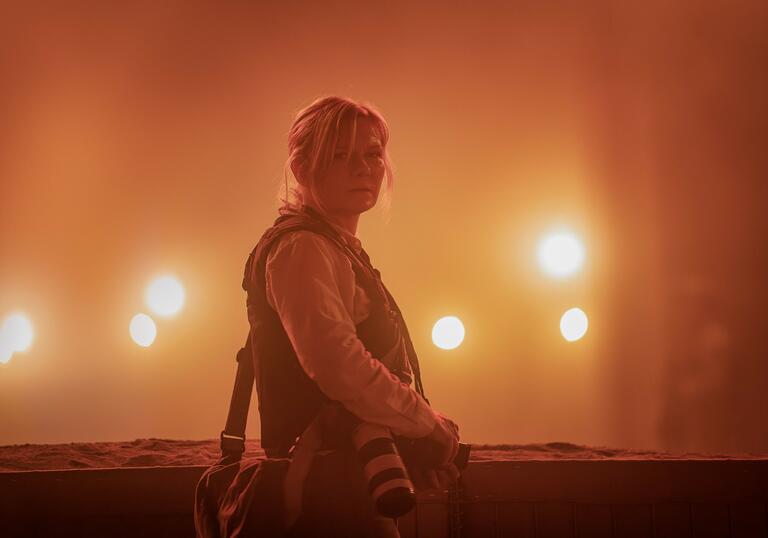 A journalist in a bullet  proof vest looks out in the night, standing in front of yellow lights.