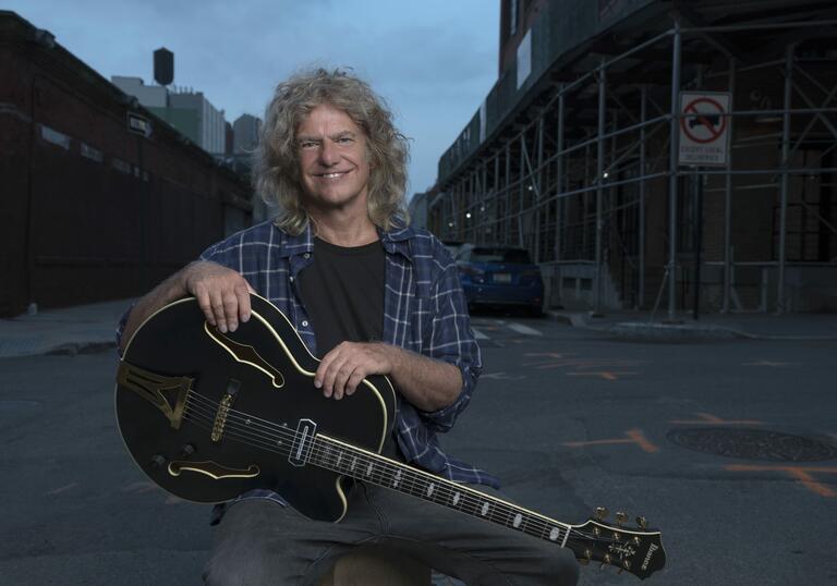 Pat Metheny sits, holding his guitar, on the street. 