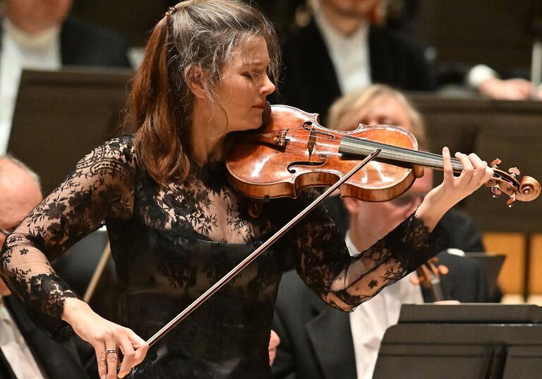 Violinist Janine Jansen performing on the Barbican Hall stage. She is wearing a laced black top and has long dark hair.