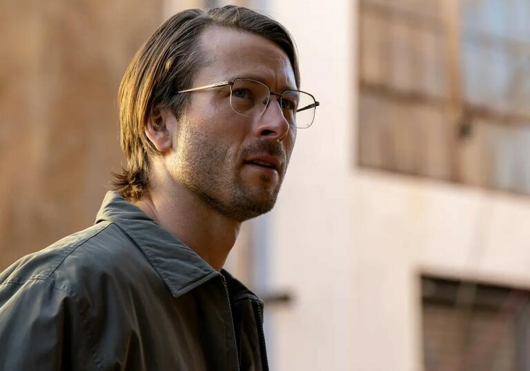 A man with slicked hair and glasses looks confused, standing on the street.