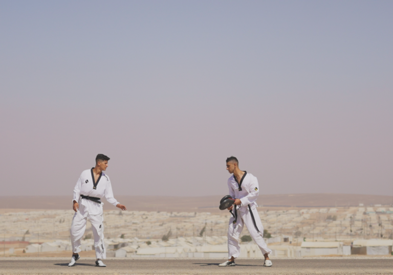 Two boys stand opposite each other wearing mixed martial arts uniforms, in front of a desert city.