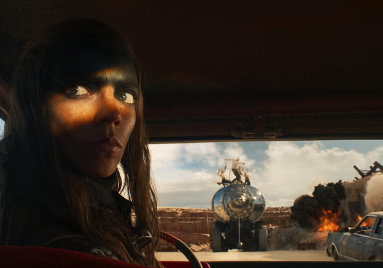 A woman in an apocalyptic landscape sits in a dark vehicle reversing.