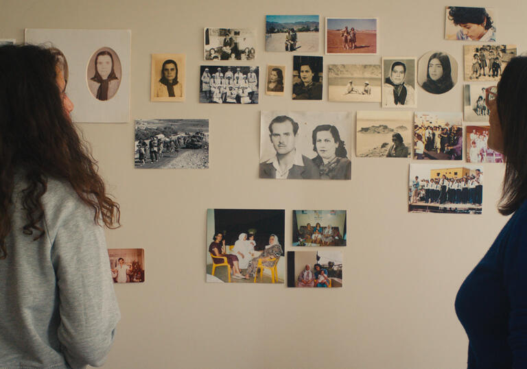 Two women look at a wall that has photographs and notes pinned on to it.
