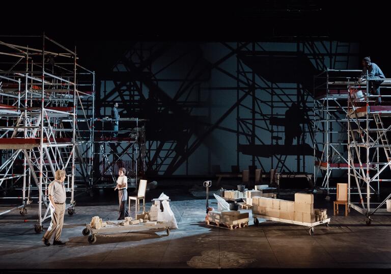 Two people stand on stage with a minimal set of furniture while tall scaffold towers line the edge of the stage.