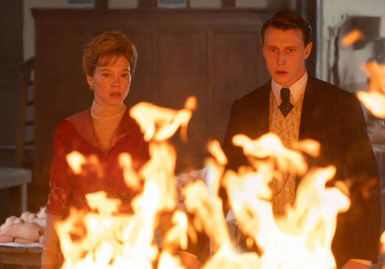 A man and a woman stand in front of a large, internal flame, dressed in a style from the 1960.