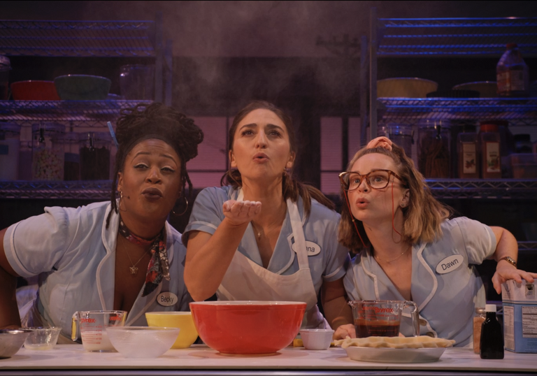 Three women in waitress outfits blow flour above a bowl.