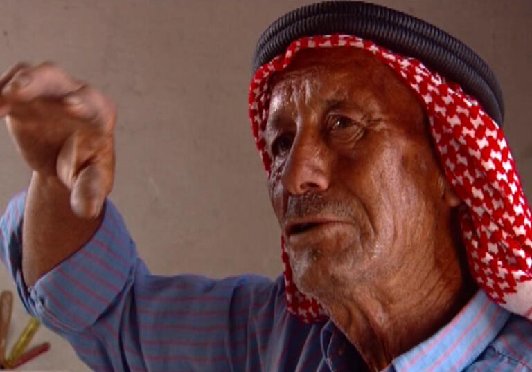 An older man in a blue shirt and head wrap talks whilst animating with his hand.