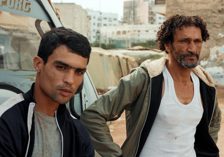 A father and son stand by a car in a parking lot in a Moroccan suburb.