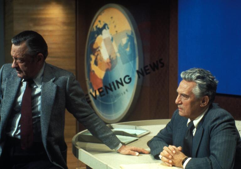 A man sits at a 70s American news broadcast desk.