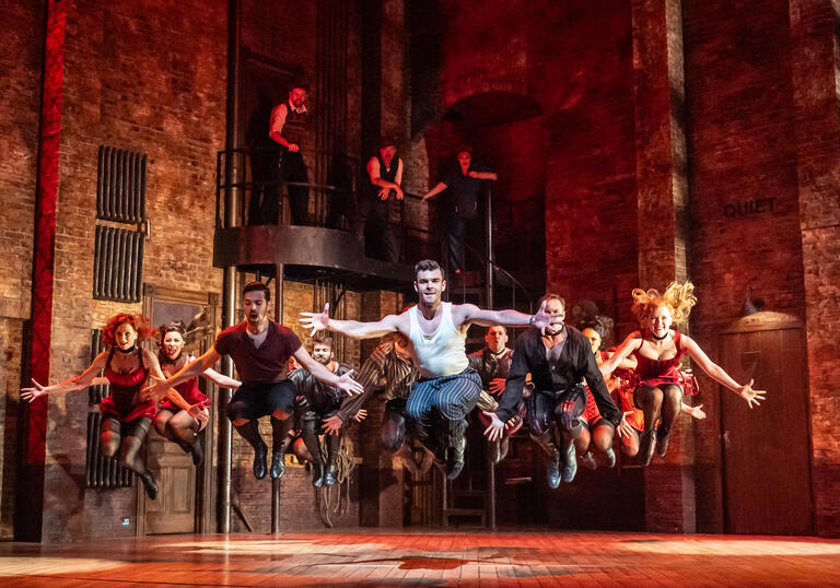 The Kiss Me, Kate ensemble perform, a group of dancers jump in the air, lit by a red light.
