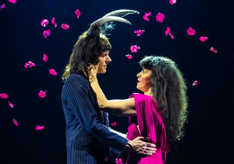 Two people embrace as pink petals fall around them. One person wears antlers on their head.