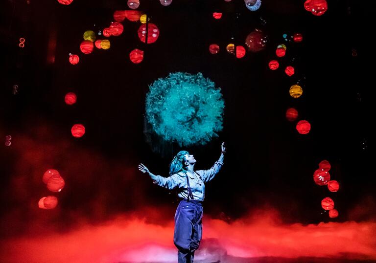 A person stands on stage looking up at a burst of aqua blue light. Red and yellow lanterns hang above her.