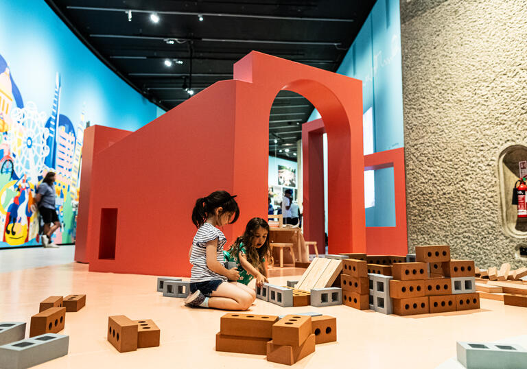 Two young girls sit on the floor in Our Street playing with some building blocks shaped like house bricks.