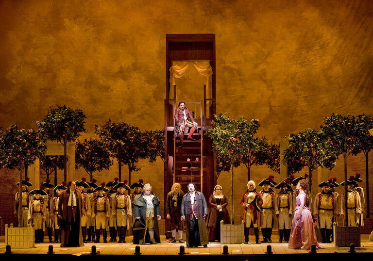 A troupe of performers stand on a golden stage surrounded by olive trees.