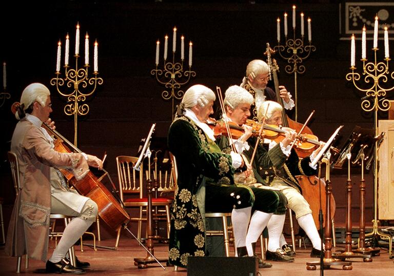 Musicians performing on stage in period costume, with five tall candelabras behind them