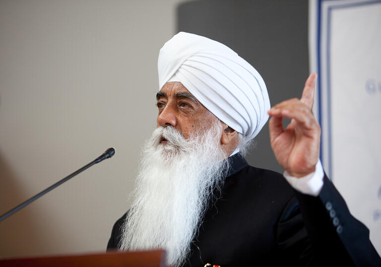 Bhai Sahib Mohinder Singh Ji speaking into a microphone, holding his finger up
