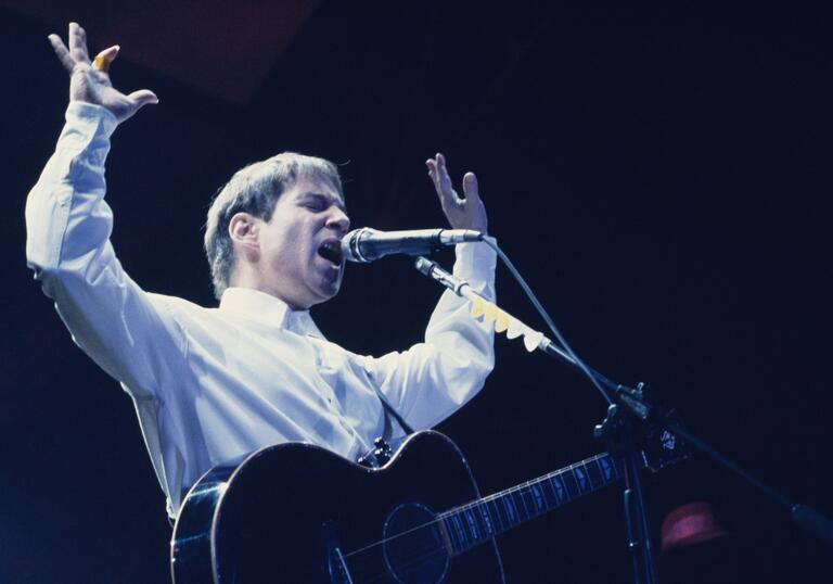 Paul Simon sings passionately on stage, a guitar around him and his hands in the air.