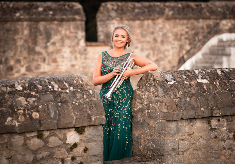 Matilda Lloyd in a green dress, holding her trumpet. She is stood within the walls of an old stone castle.