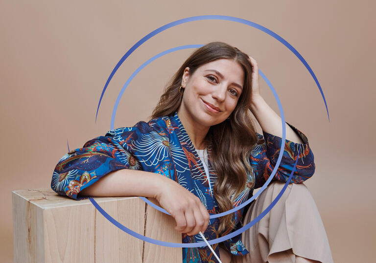 Dalia Stasevska sitting down, resting her arm on a wooden block. There are blue circular swirls around her.