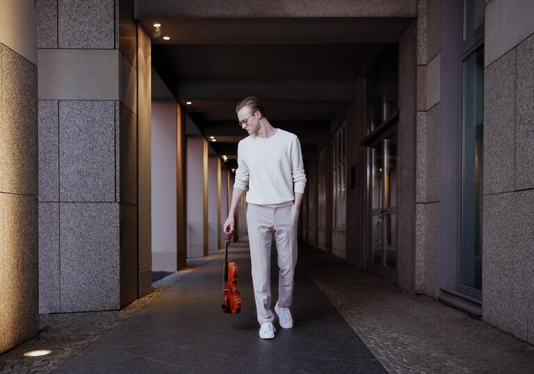 Sao Soulez Lariviere waking down a concrete corridor holding his viola by his side