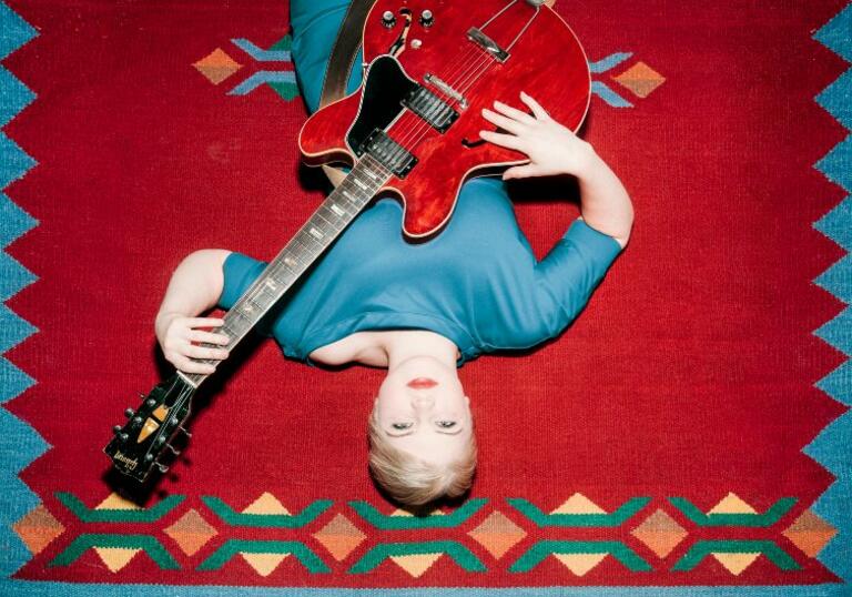 LILJA lays on a red and blue carpet, holding a red guitar