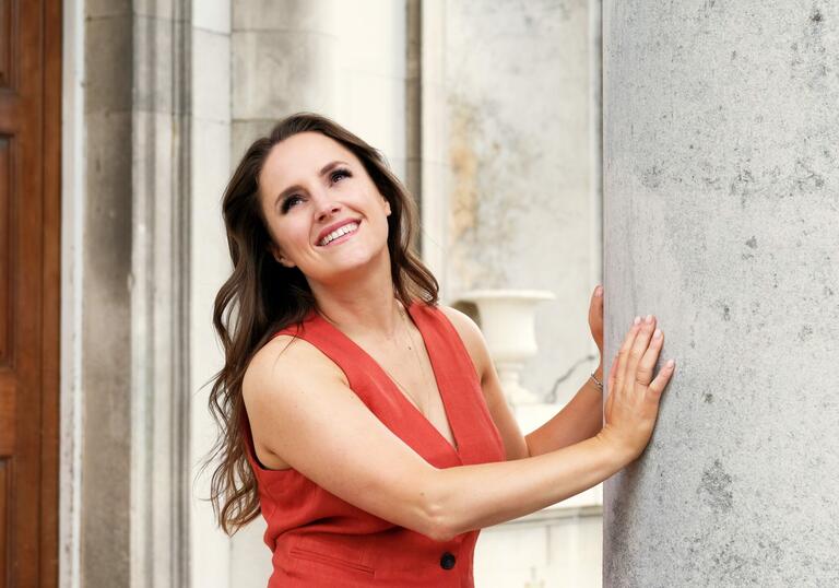Louise Alder standing by a large concrete pillar, smiling and looking upwards