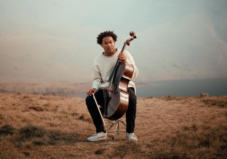Sheku Kanneh-Mason sitting with his cello in a mountainous landscape, with a lake in the distance behind him