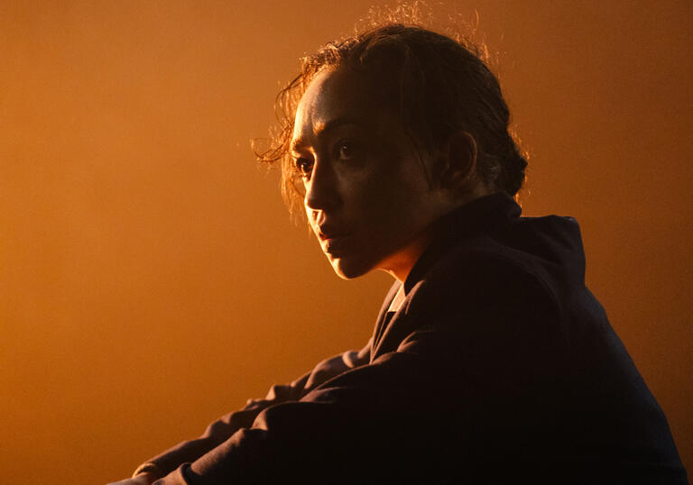 Actor Ruth Negga stares thoughtfully off-camera as a golden glow lights the stage behind her.