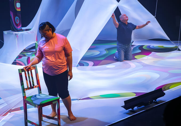Two people perform on stage among large pieces of draping white fabric with bright lights projected onto it.