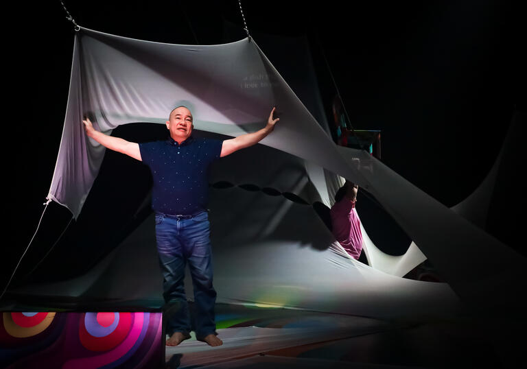A person performs underneath a large piece of white draping fabric, they hold their arms out wide.
