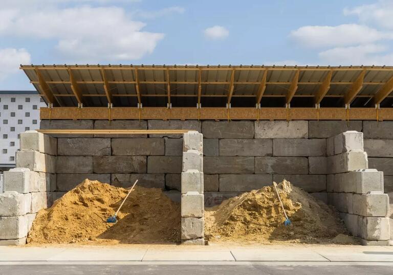 A building in construction, with cement blocks forming the frame and a partial timber frame roof. There are large piles of sand on the floor.