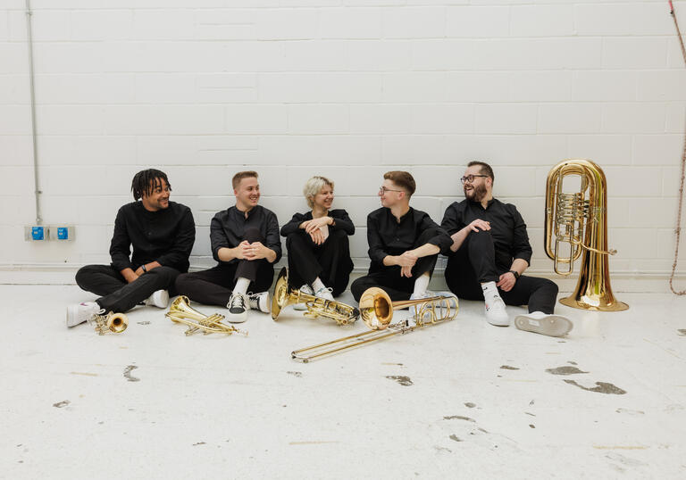 The members of Connaught Brass sitting in a row in front of a white wall, their instruments placed on the floor in front of them.