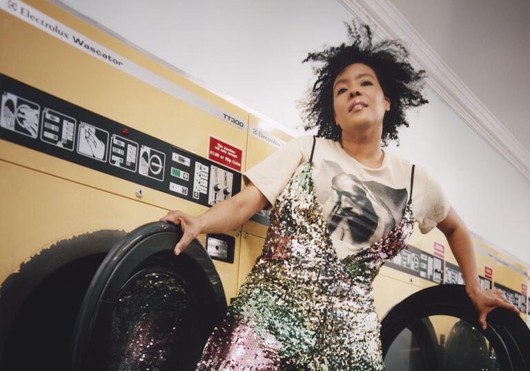 Elaine Mitchener standing in front of washing machines in a laundrette, holding the open washing machine doors.