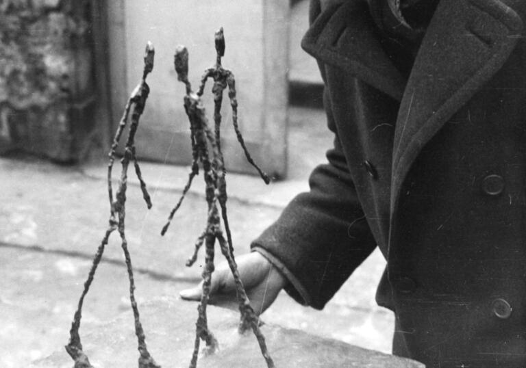 A black and white photo of Alberto Giacometti in his studio holding his sculpture, Three Men Walking.