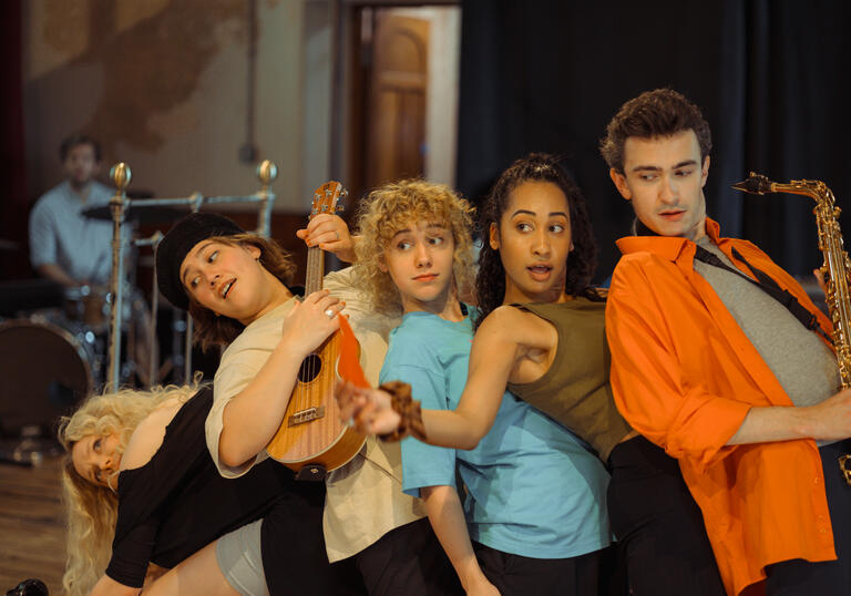 Five performers lean on each other in a line. Two play instruments.
