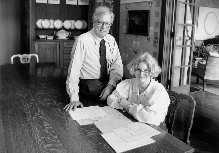 Black and white photo of Robert Venturi and Denise Scott Brown. She is sat at a large table and he is next to her, perched on the table.
