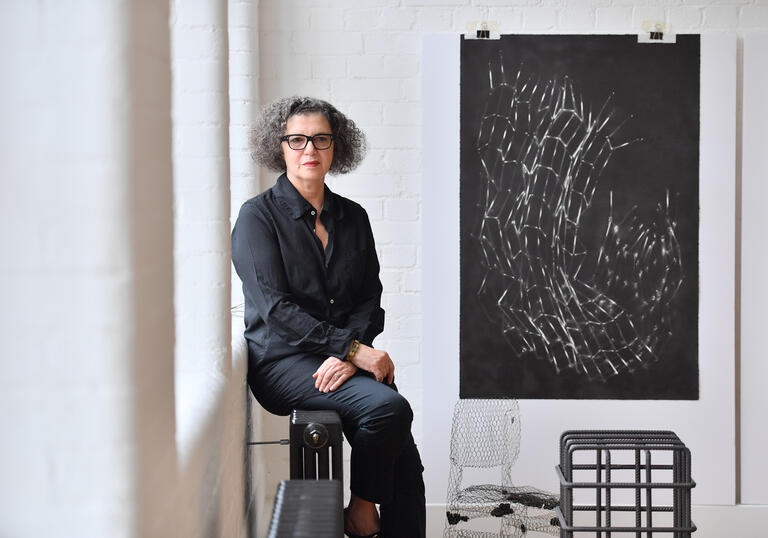 Mona Hatoum sitting on a radiator on the wall of her studio. On the wall next to her is Untitled (Bed Springs), one of her lithograph print depicting a structure made of bed springs, curved and crumpled.