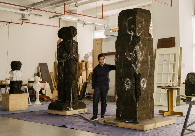Huma Bhabha standing in a studio with two tall sculptures, one carved out and one with markings ready to be carved.