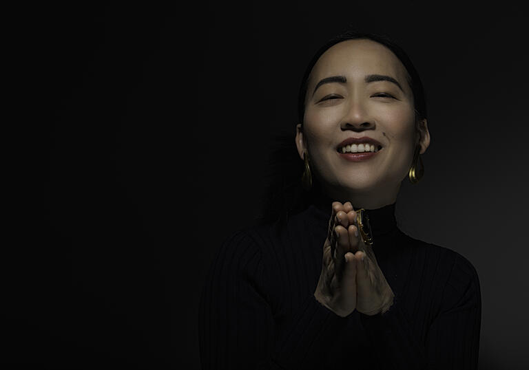Helen Sung against black background, smiling with eyes closed and hands in prayer position