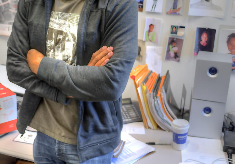 A man stands leaning against a desk. The wall behind is filled with images.