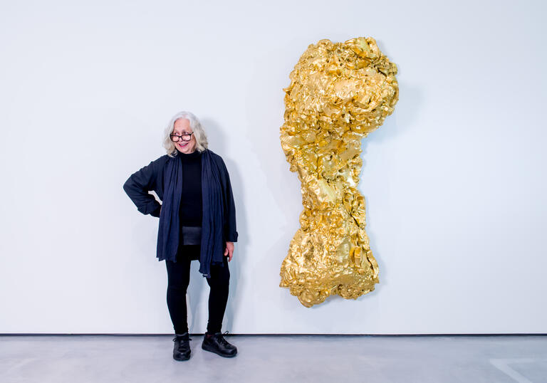 Lynda Benglis standing next to one her sculpture titled Ghost Dance/Pedmarks. It is a large, gold, torso-like form hanging on a white wall.