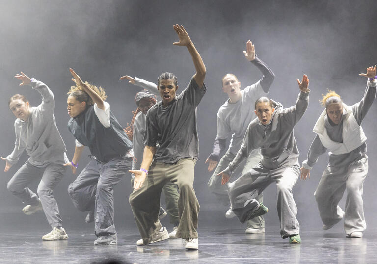 A group of hip hop dancers caught in motion. They lift one arm in the air and open their mouths.