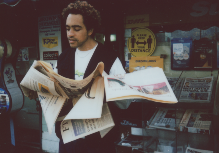 A person stands outside a newsstand, reading a newspaper. Signs and various magazines are visible in the background.