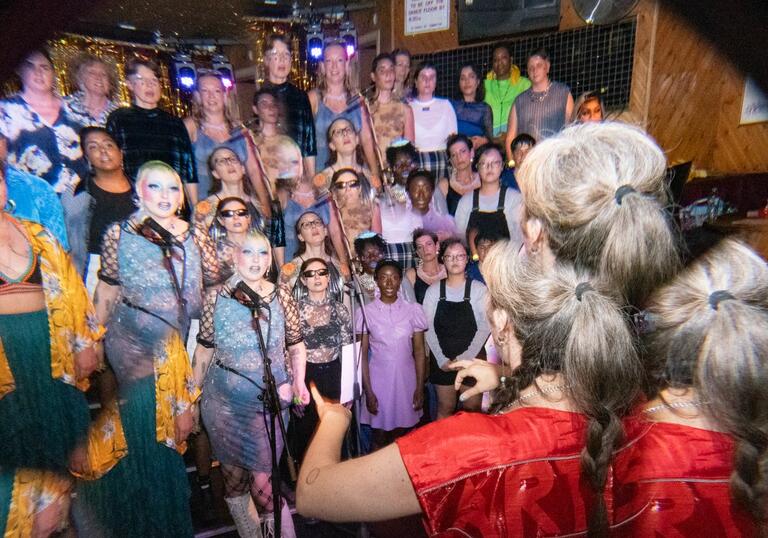 A choir group stands on stage in a dimly lit venue, with diverse singers wearing colorful outfits
