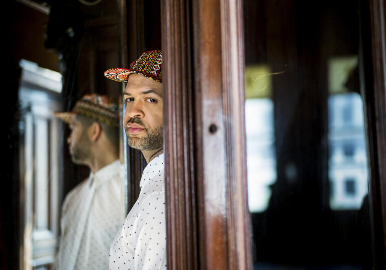 Jason Moran standing in a doorway. The back half of his body is obscured by the door frame, and he is turning to face the camera. His reflection is visible in a mirror behind him.