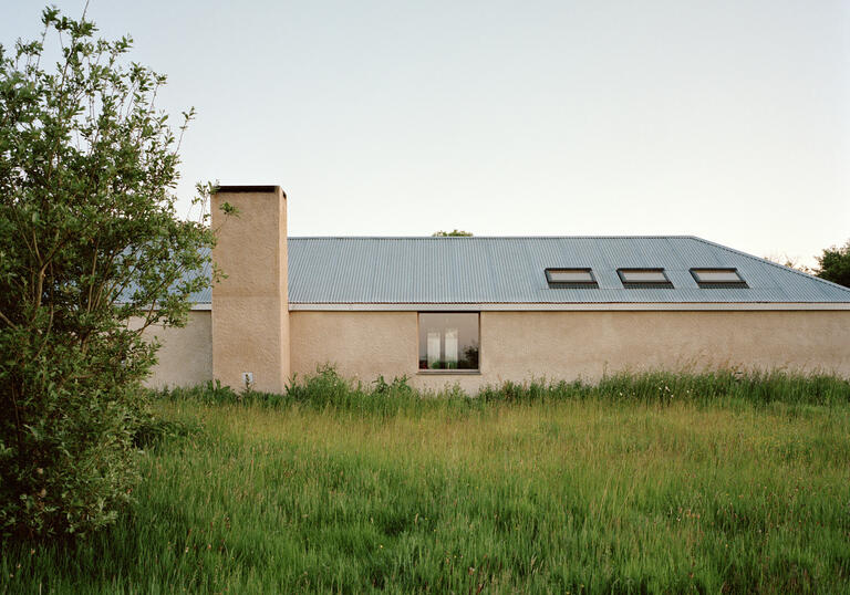 A photo of the Farmworker's House in Cornwall. It is a single storey, with a lime render and a pitched roof with three roof windows on the right side. On the left is a large chimney. It is surrounded by a large meadow of grass.