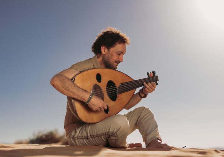 Portrait of Naseer Shamma sitting on sand and playing the oud